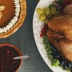 A neatly laid out Thanksgiving dinner table, featuring a golden-brown turkey, pumpkin pie and cranberry sauce on a grey tablecloth.