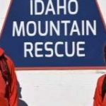 Four people in red jackets standing in front of a blue sign shaped like a mountain with the words Idaho Mountain Rescue on it.