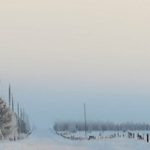 A long snow-covered country road with power poles along one side and a barn in the distance.